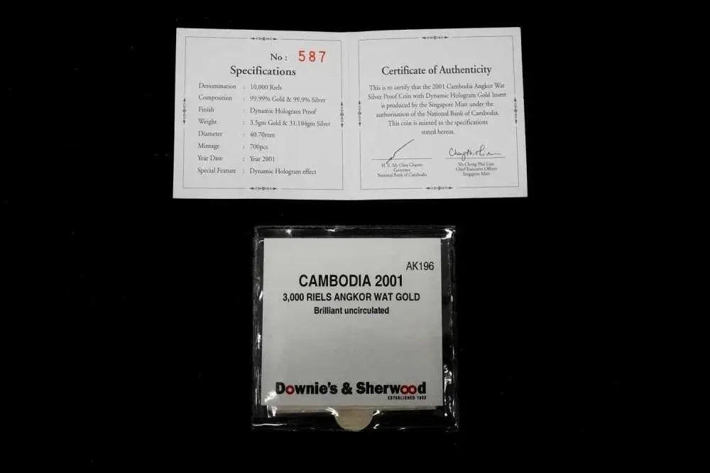 A packaged gold coin from Cambodia, 3,000 Reils Angkor Wat Gold, shown below its authenticity certificate and specifications card on a black background. The coin is sealed in a clear plastic bag with a white label, and the certificate is printed on white paper.