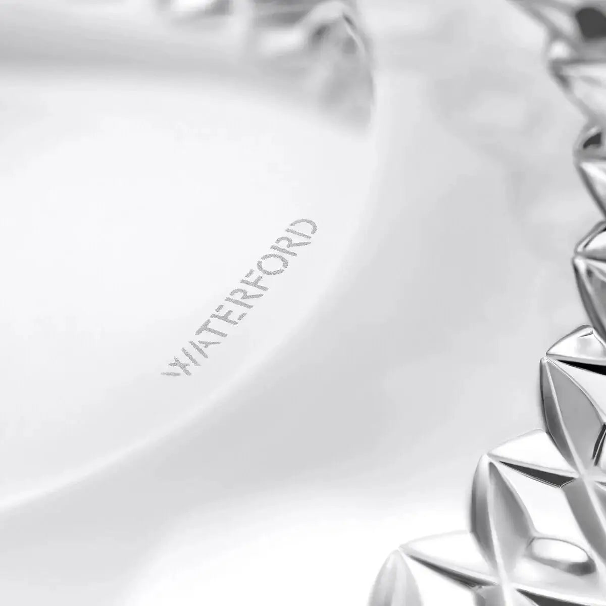 Close-up of a clear glass plate with the brand name 'WATERFORD' etched on the inside, showing a faceted silver-toned glass rim on the right side.