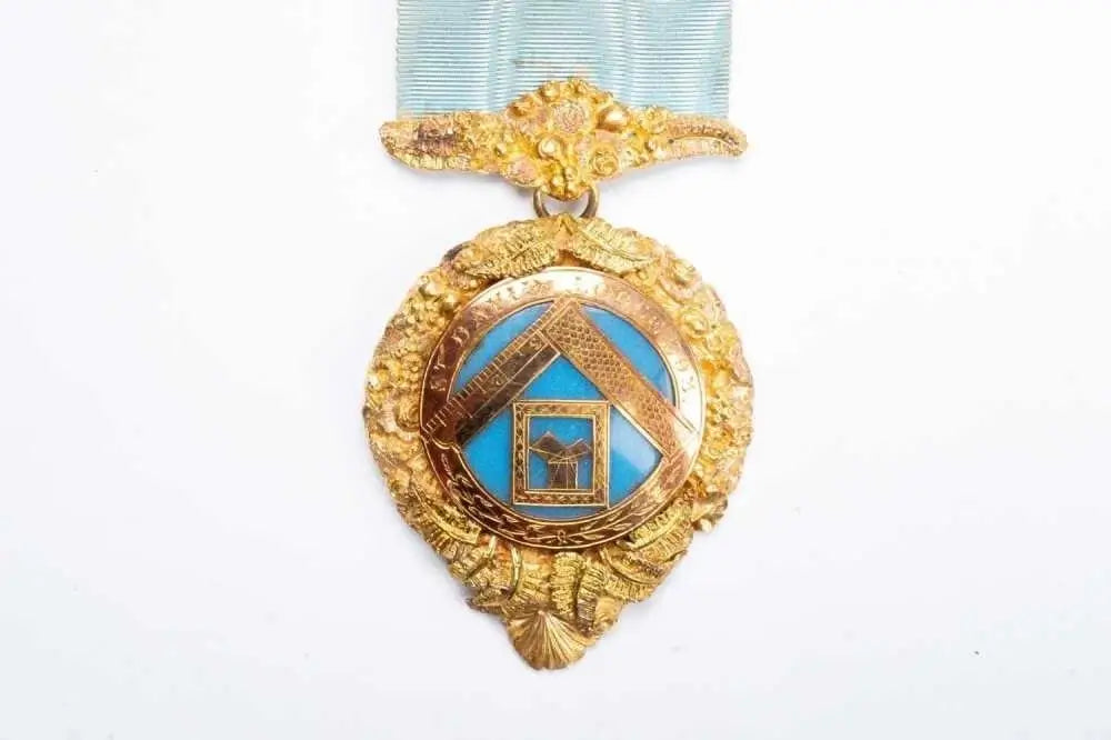 A gold-toned Masonic square and compass pendant medal with light blue enamel details, hanging from a light blue ribbon on a plain white background.