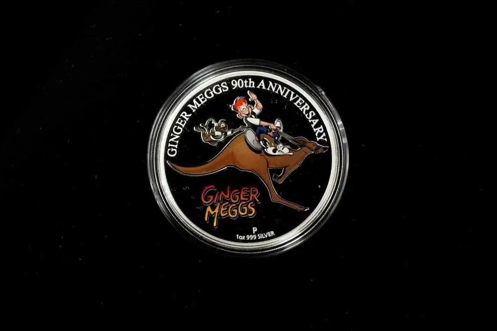 A round silver commemorative coin or medallion on a black background, featuring an illustration of a person riding a kangaroo with the text 'GINGER MEGGS 90TH ANNIVERSARY' and 'GINGER MEGGS'.
