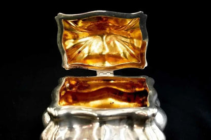 A close-up of an ornate silver-toned jewelry box with its lid open, showing a reflective amber-gold interior against a black background.