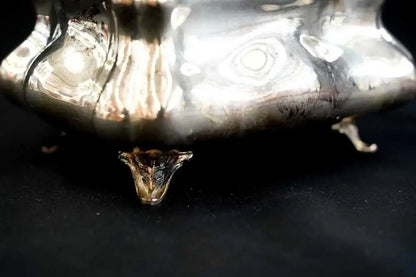 A close-up of a polished silver decorative object with three small claw feet, photographed against a dark black background.