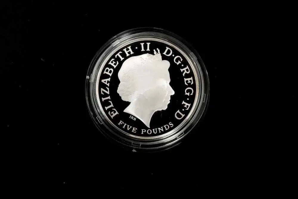 A silver five-pound coin featuring the profile of Queen Elizabeth II, encased in a clear protective plastic capsule, set against a solid black background.