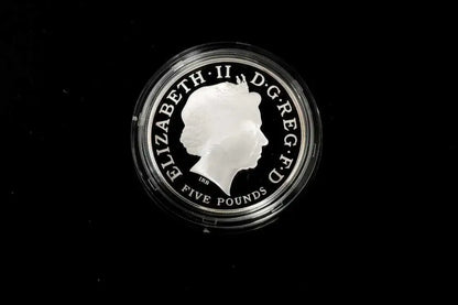 A silver five-pound coin featuring the profile of Queen Elizabeth II, encased in a clear protective plastic capsule, set against a solid black background.