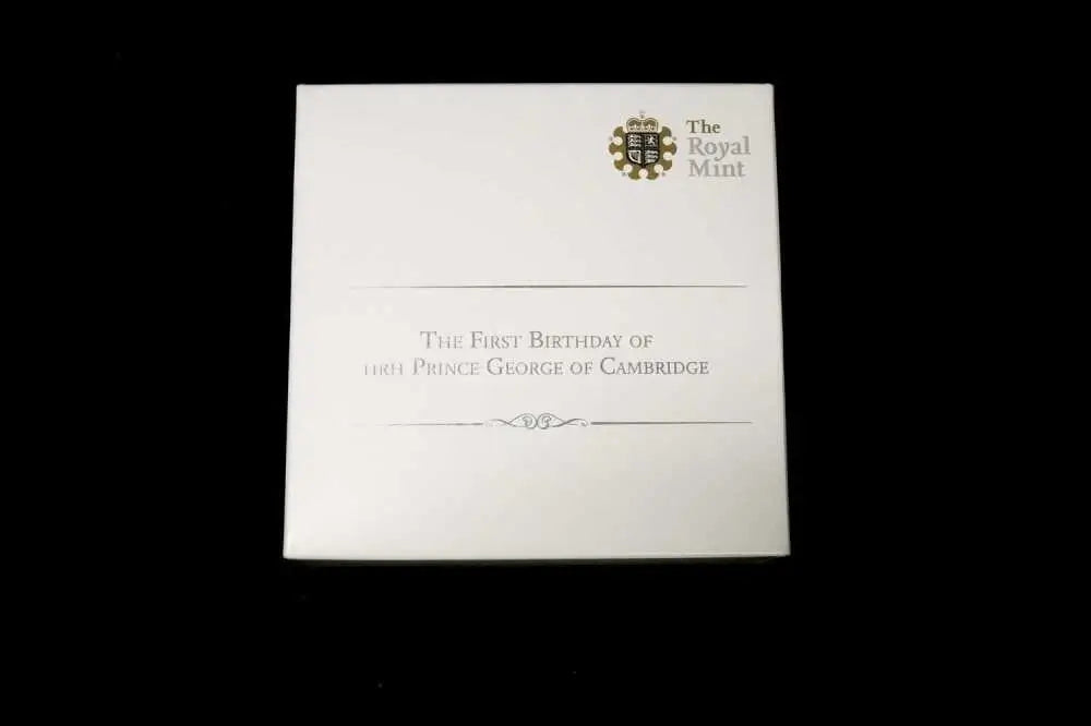 A square white card or box from The Royal Mint, featuring black text that reads 'The First Birthday of Her Royal Majesty's Prince George of Cambridge' with a small crest at the top right, shown against a solid black background.