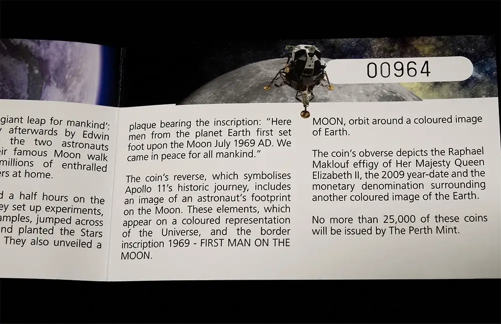 A close-up of a coin or collectible plaque insert with printed text about moon exploration and an astronaut orbit illustration. The visible text includes 'MOON, orbit around a coloured image of Earth.