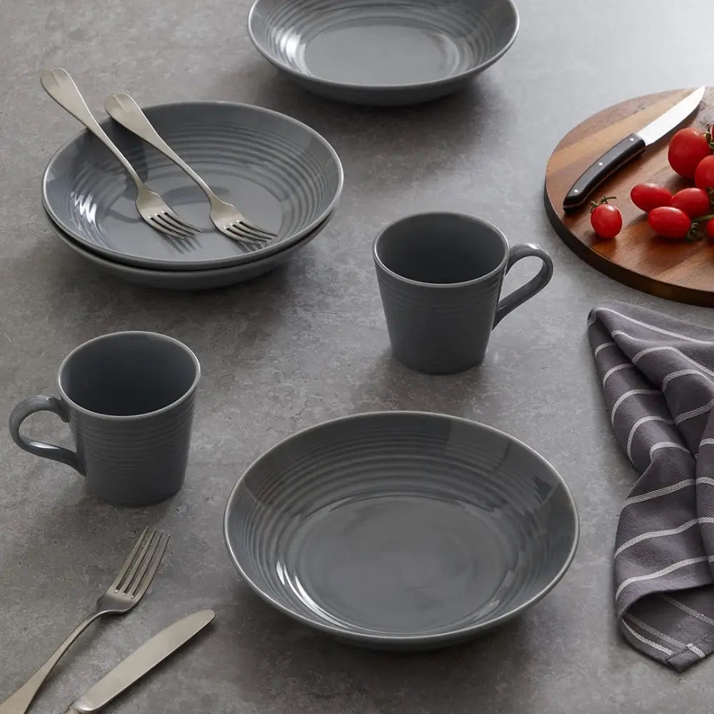 Gray ceramic dinnerware set on a gray surface with cutlery and a cutting board.