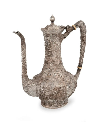 An ornate silver pewter coffee pot with intricate floral engravings, a long curved spout, and a decorative handle, shown on a white background.