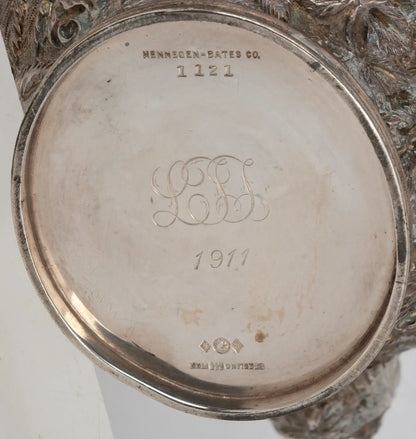 Close-up of the bottom of an ornate silver-toned metal object, showing engraved text including 'MENNEGEN - BATES CO.', '1121', a monogram 'LBF', and '1911'.