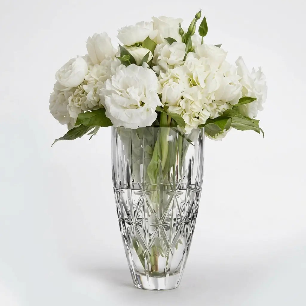 A bouquet of white flowers with green leaves arranged in a clear cut-crystal vase against a light gray studio background.