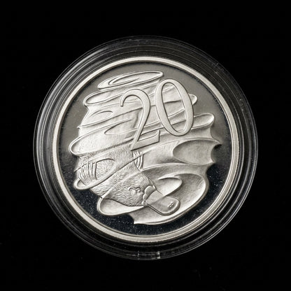 A circular silver coin or medallion inside a clear protective plastic capsule, photographed against a solid black background. The coin features raised abstract wave-like forms and the number 20, with a small bird motif near the bottom.