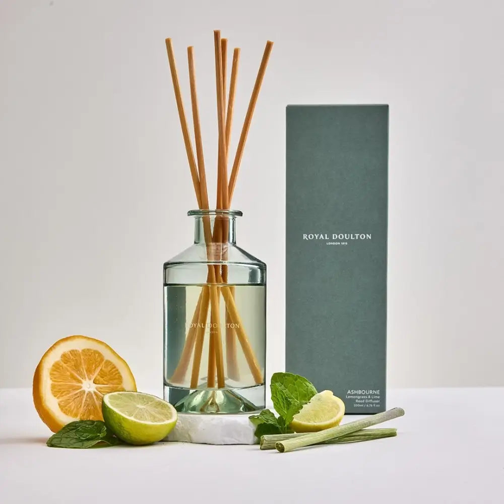 Reed diffuser with wooden sticks, citrus fruits, and mint leaves on a white surface with a Royal Doulton box.