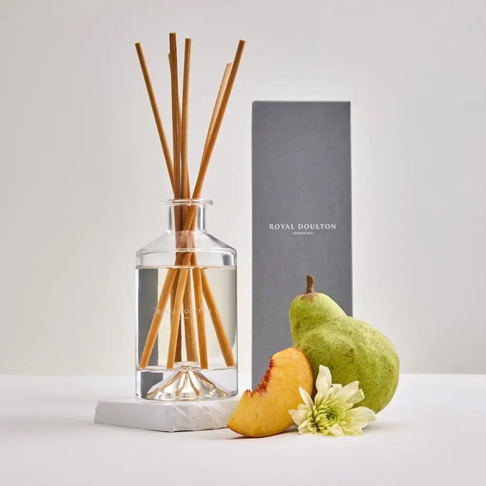 Diffuser with wooden sticks, pear, peach, and flower on a white surface with a gray box in the background.