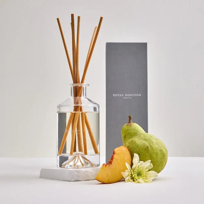 Diffuser with wooden sticks, pear, peach, and flower on a white surface with a gray box in the background.