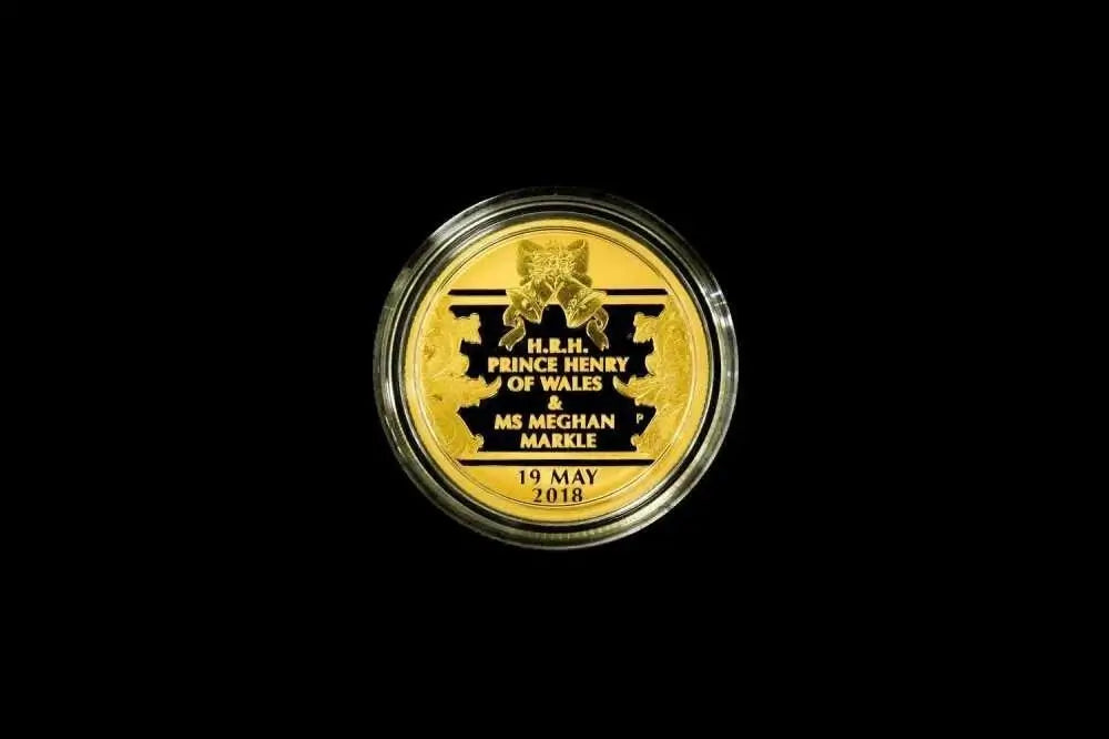 A round gold commemorative coin in a clear protective capsule, centered on a solid black background. The coin features black text and a black crest with the text 'H.