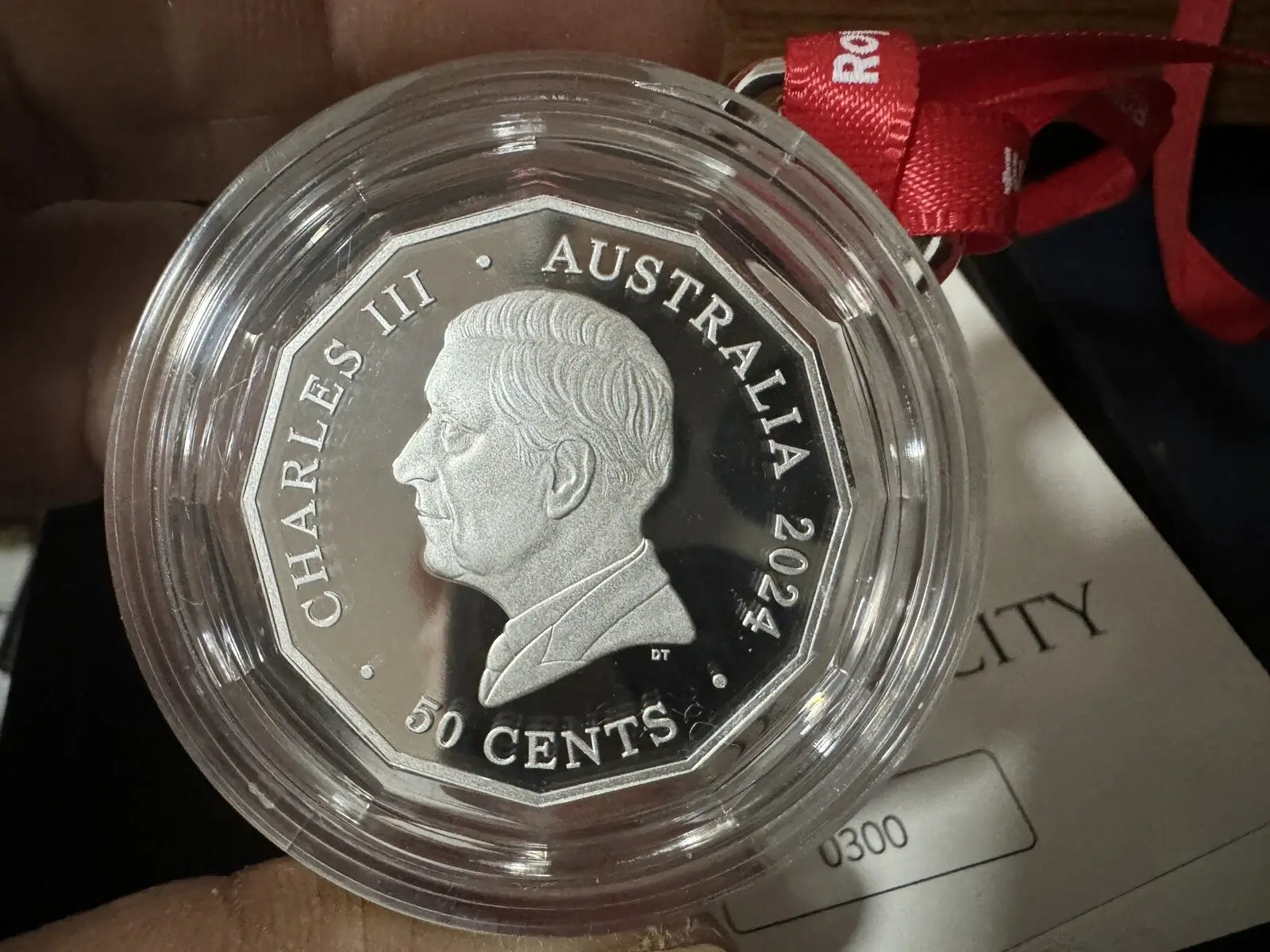 A 2024 Australia 50 cents coin in a clear protective capsule, held by a hand with a red ribbon tag attached, next to a Certificate of Authenticity card.