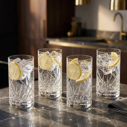 Luxury lifestyle photography of a set of 4 Royal Doulton Harper Highball glasses filled with sparkling water, ice, and lemon slices, arranged on a dark marble surface with cinematic lighting.