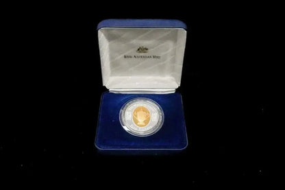 A collectible coin presented in an open blue velvet case against a black background. The coin is housed in a clear circular holder, with a gold-toned inner ring, and the case lid is lined with white satin featuring the text 'ROYAL AUSTRALIAN Mint'.