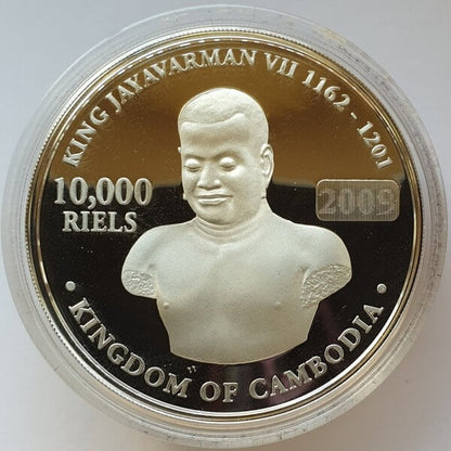 A round commemorative coin of Cambodia, featuring King Jayavarman VII, displayed inside a clear protective plastic capsule. The coin has a gold and black background with silver-toned relief details and text reading '10,000 RIELS' and '2009'.
