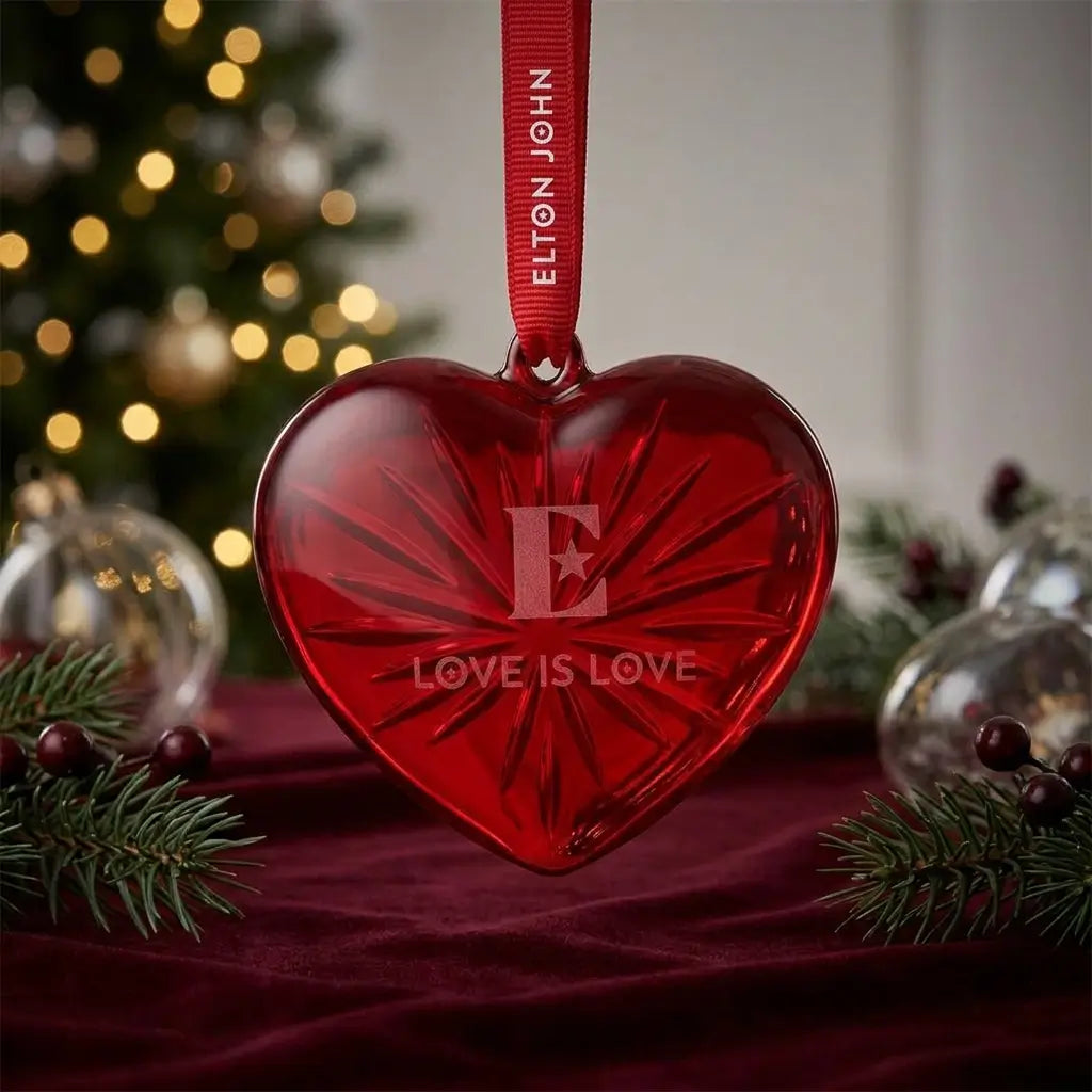 Red heart-shaped ornament with 'Elton John' and 'Love is Love' text, Christmas tree in the background.