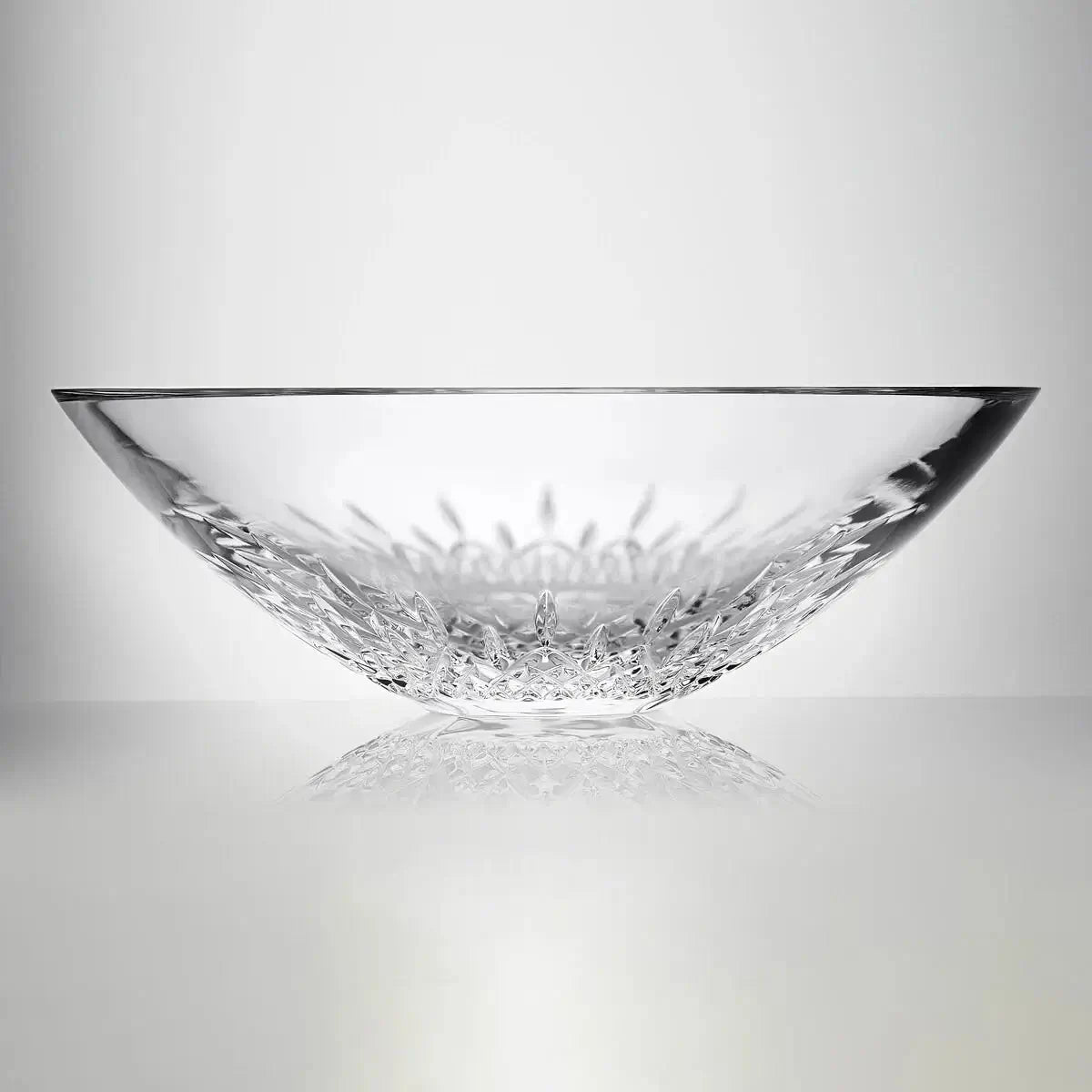 A clear cut-glass bowl with a textured, faceted base, photographed on a reflective light gray surface against a neutral gray background.