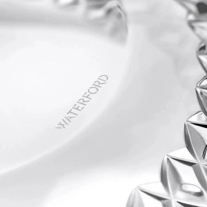 Close-up of a clear glass plate with the brand name 'WATERFORD' etched on the inside, showing a faceted silver-toned glass rim on the right side.