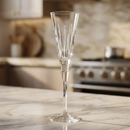 A clear cut-crystal champagne flute with vertical faceted detailing, standing on a light marble countertop with a softly blurred kitchen background.