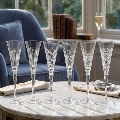 Six clear cut-crystal champagne flutes arranged on a round marble table in a bright living room, with a blue armchair, books, and a window in the background from our Waterford Crystal Australia – Outlet & Sale Collection