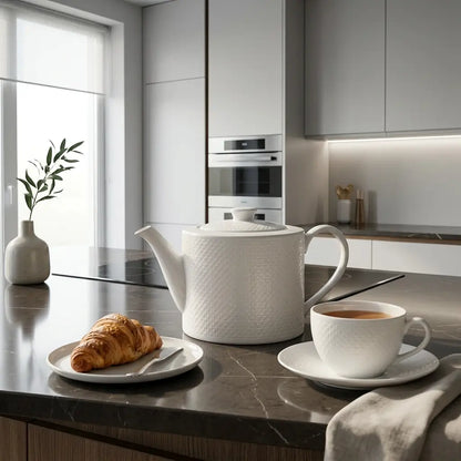 White teapot and cup on a kitchen counter with a croissant and coffee.