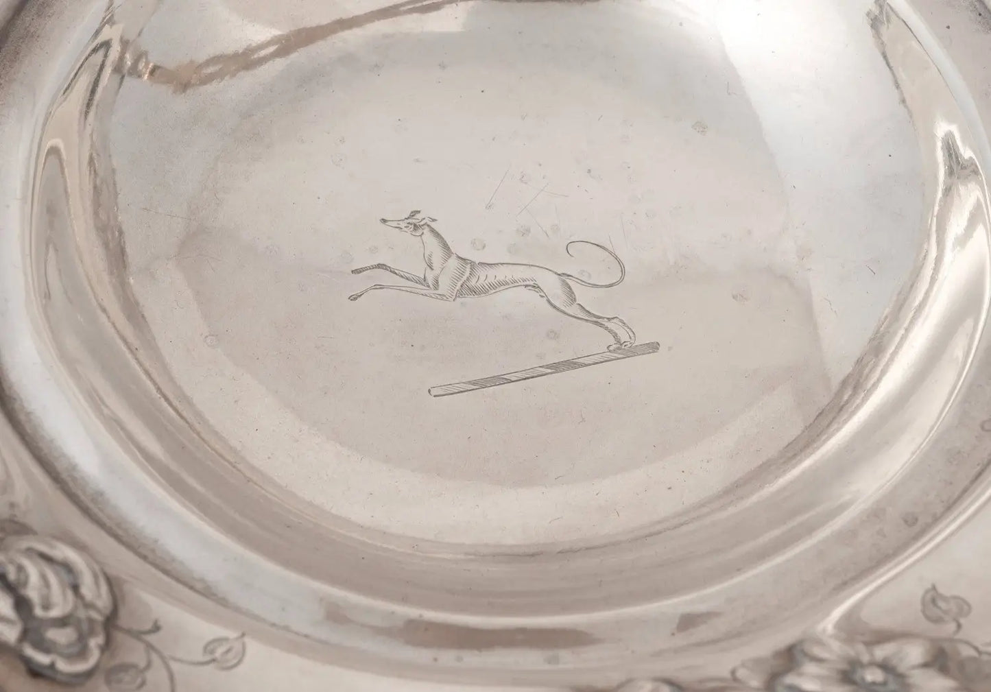 Close-up of the interior of a polished silver-toned bowl or plate, showing a engraved leaping dog motif near the center.