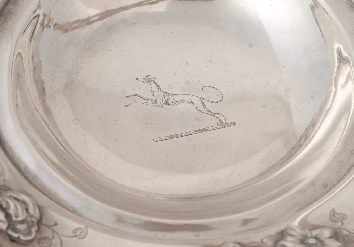 Close-up of the interior of a polished silver-toned bowl or plate, showing a engraved leaping dog motif near the center.