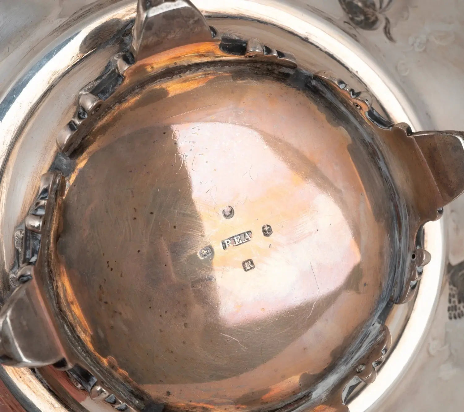 Close-up of the interior bottom of a polished silver-toned metal pot or vessel, showing a reflective interior surface with a small maker's mark and decorative edge details.