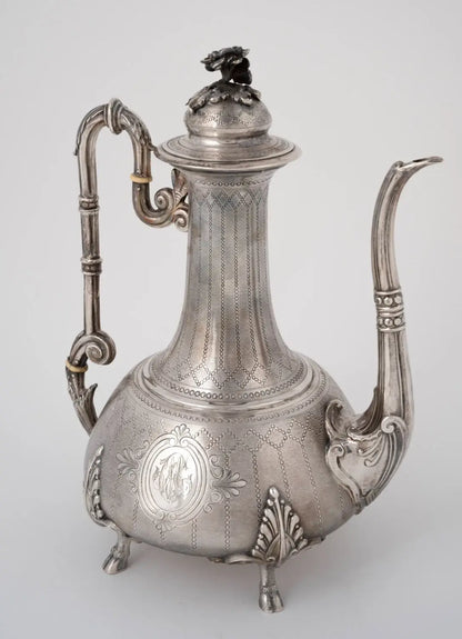 An ornate silver coffee pot with a decorative handle, curved spout, and lid topped with a small lion figurine, photographed against a light gray background.