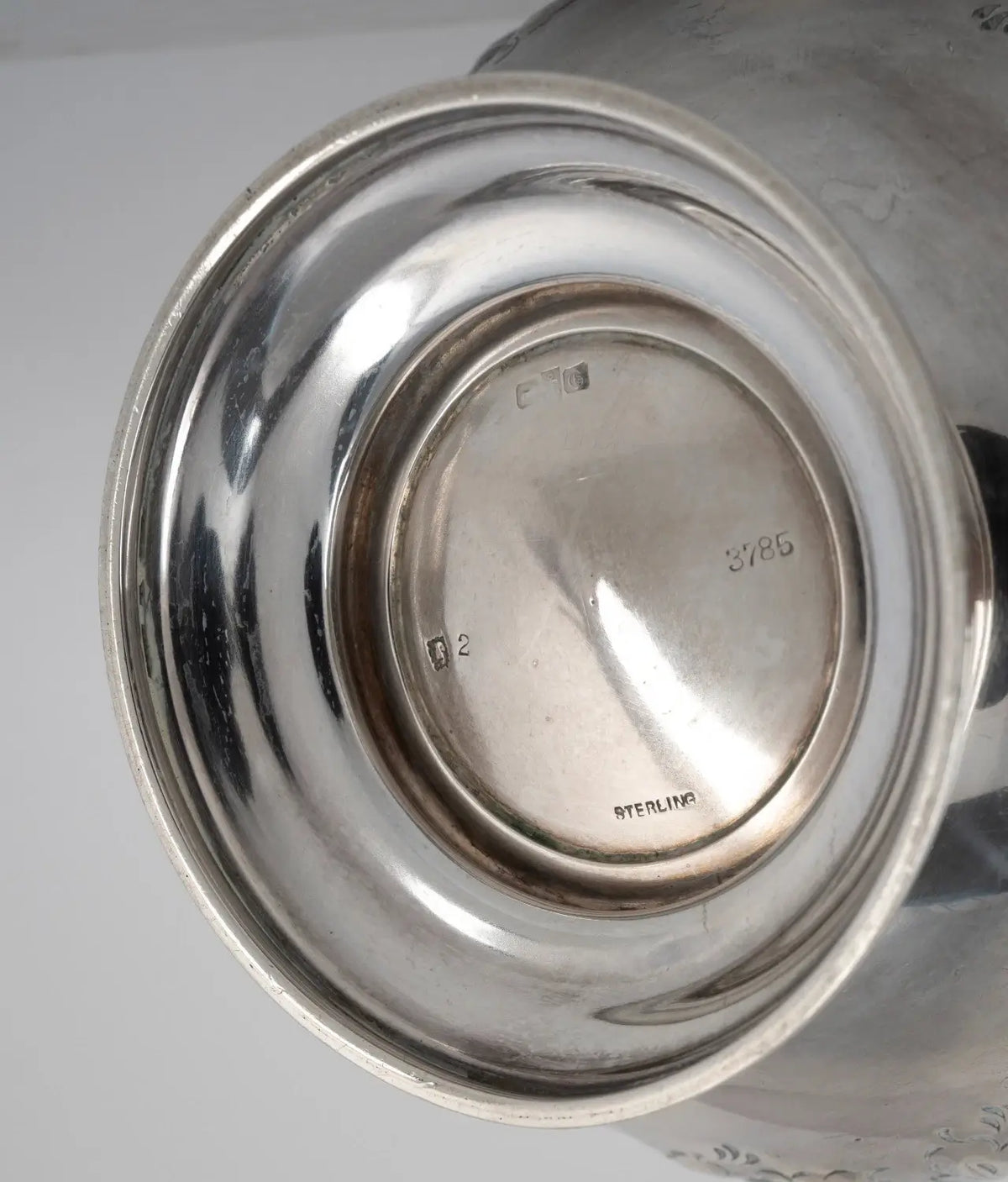 Close-up of the polished silver base of a metal vessel, showing engraved maker's marks and the word 'STERLING' on a light gray background.