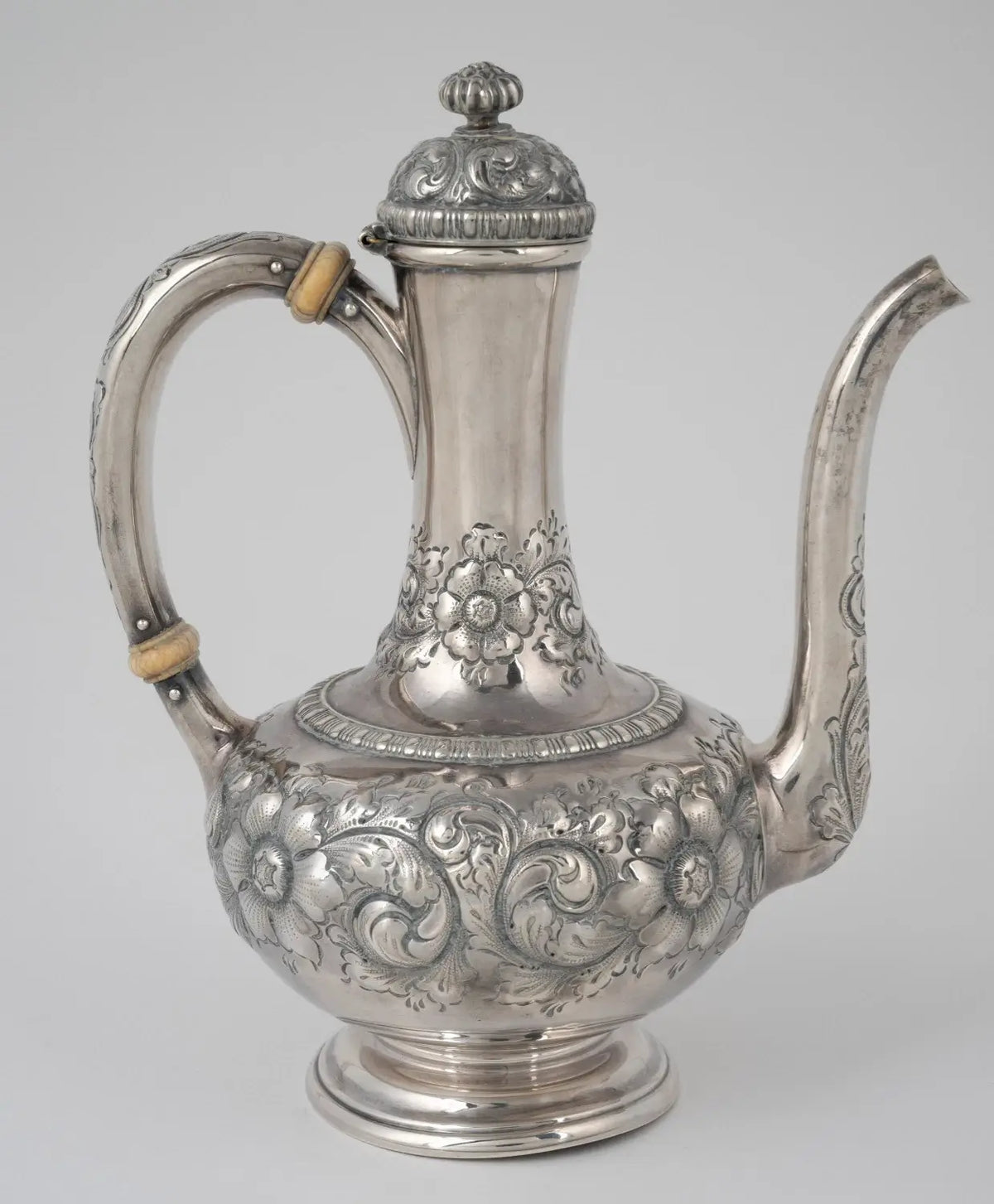 An ornate silver coffee or tea pitcher with intricate floral and scroll engravings, featuring a curved handle with light wood accents and a long spout, photographed against a neutral gray background.