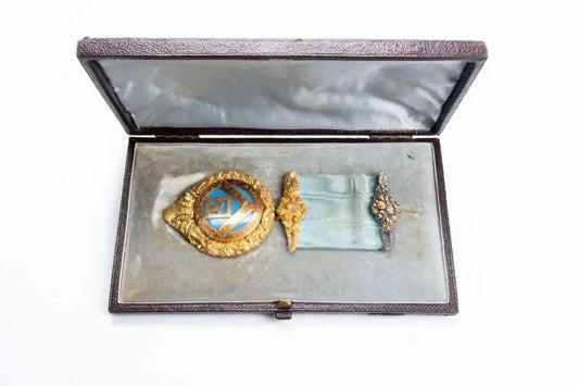 A vintage Masonic square and compass badge with a blue and gold center, displayed inside an open dark brown presentation case with a gray velvet lining.