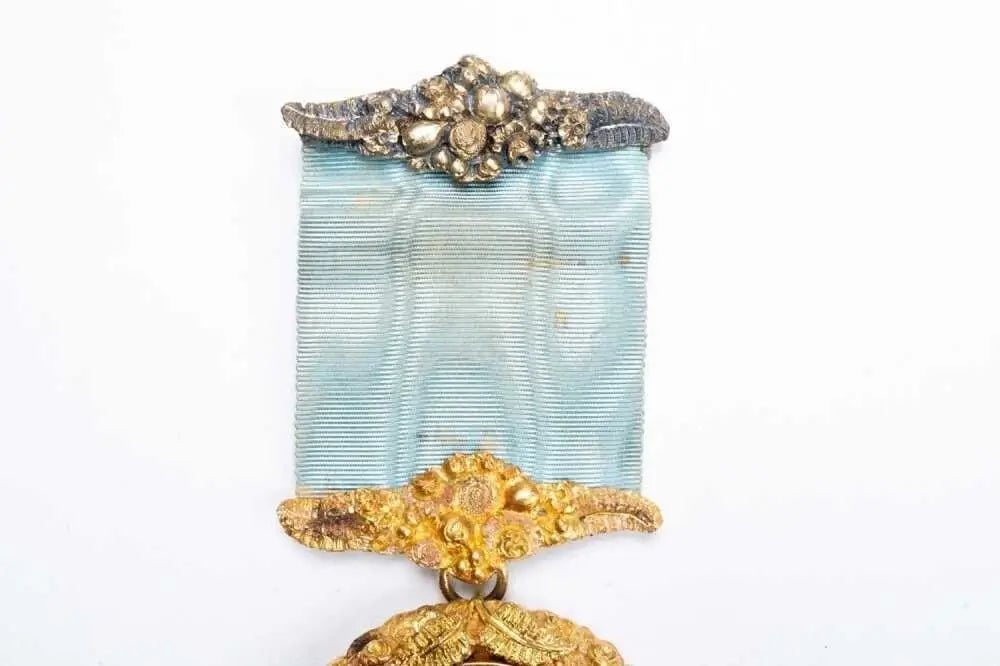A vintage medal featuring a light blue ribbon with ornate gold-toned metal detailing at the top and bottom, shown against a plain white background.