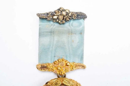 A vintage medal featuring a light blue ribbon with ornate gold-toned metal detailing at the top and bottom, shown against a plain white background.