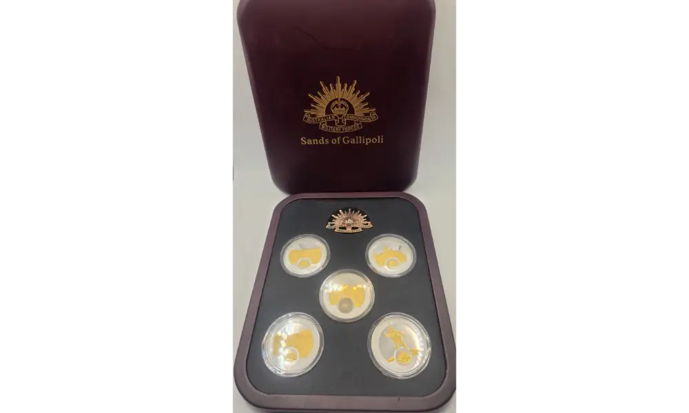 A set of five gold-plated coins displayed in a black tray with a dark reddish-brown presentation box behind it, featuring the 'Sands of Calipoli' logo.
