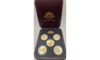 A set of five gold-plated coins displayed in a black tray with a dark reddish-brown presentation box behind it, featuring the 'Sands of Calipoli' logo.
