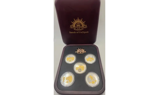 A set of five gold-plated coins displayed in a black tray with a dark reddish-brown presentation box behind it, featuring the 'Sands of Calipoli' logo.