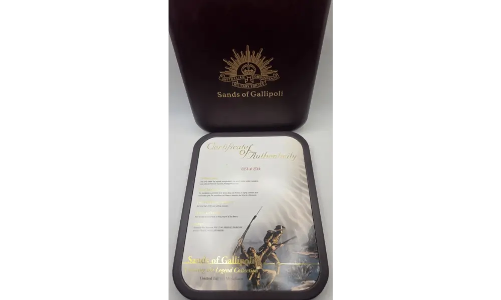A dark reddish-brown presentation box with a gold crest and the text 'Sands of Gallipoli' sits behind a smaller matching tray or plaque featuring a printed certificate and a small figurine, on a white background.