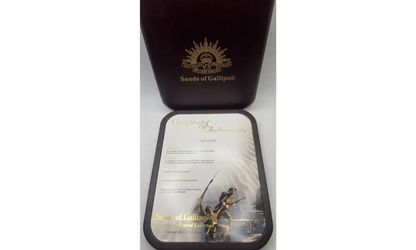 A dark reddish-brown presentation box with a gold crest and the text 'Sands of Gallipoli' sits behind a smaller matching tray or plaque featuring a printed certificate and a small figurine, on a white background.