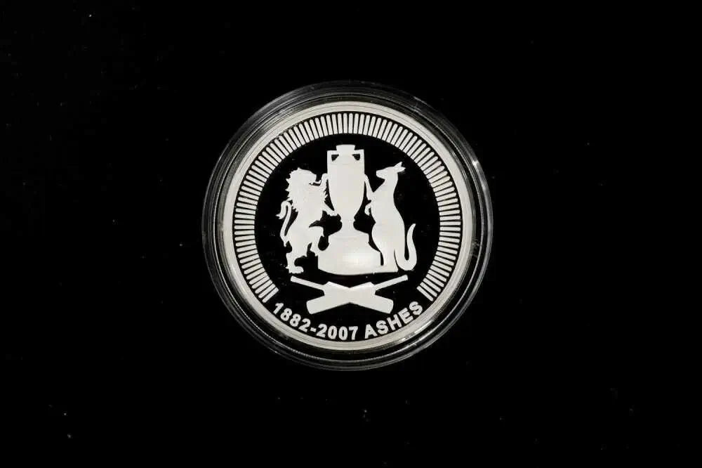 A round silver commemorative coin featuring the Ashes cricket trophy and lions and a cat, with the text '1882-2007 ASHES' on a black background.