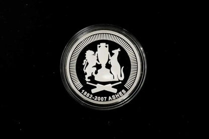 A round silver commemorative coin featuring the Ashes cricket trophy and lions and a cat, with the text '1882-2007 ASHES' on a black background.
