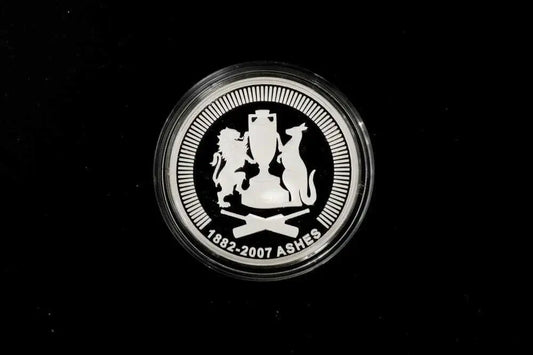 A round silver commemorative coin featuring the Ashes cricket trophy and lions and a cat, with the text '1882-2007 ASHES' on a black background.