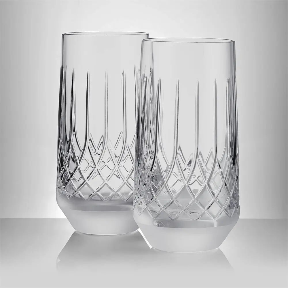 Two clear cut-crystal tumblers with vertical and diamond-pattern engravings, standing on a reflective light gray surface against a neutral gray background.