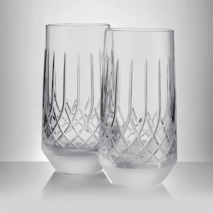 Two clear cut-crystal tumblers with vertical and diamond-pattern engravings, standing on a reflective light gray surface against a neutral gray background.