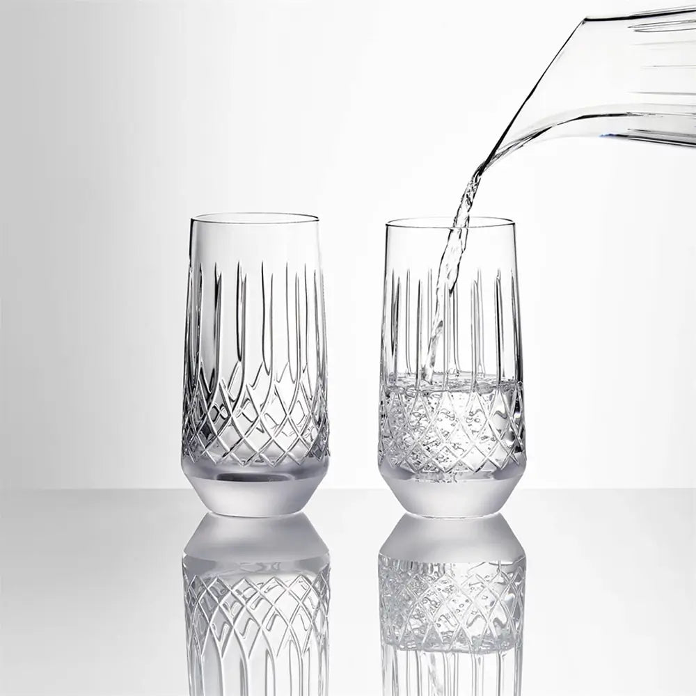 Two clear cut-crystal glasses on a reflective white surface, with water being poured into one glass from a transparent pitcher against a light gray background.