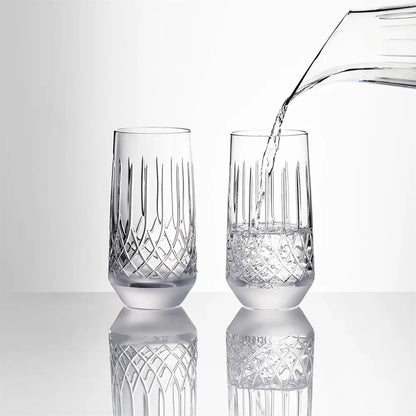 Two clear cut-crystal glasses on a reflective white surface, with water being poured into one glass from a transparent pitcher against a light gray background.